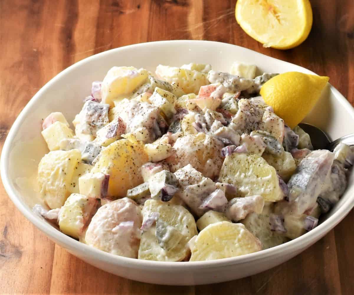 Side view of potato herring salad in white bowl with lemon in background.
