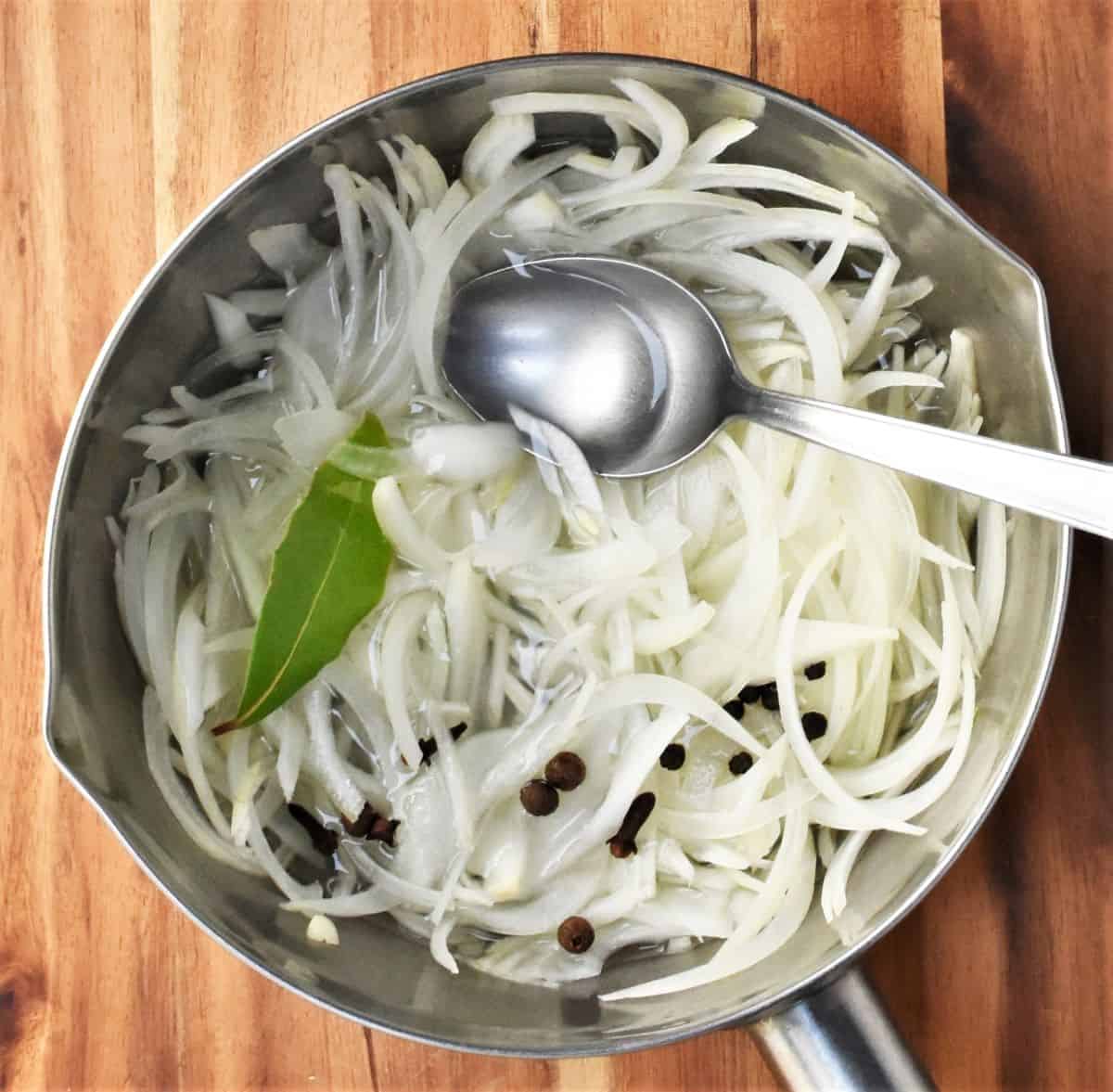 Slices of onion and spices in saucepan with spoon.