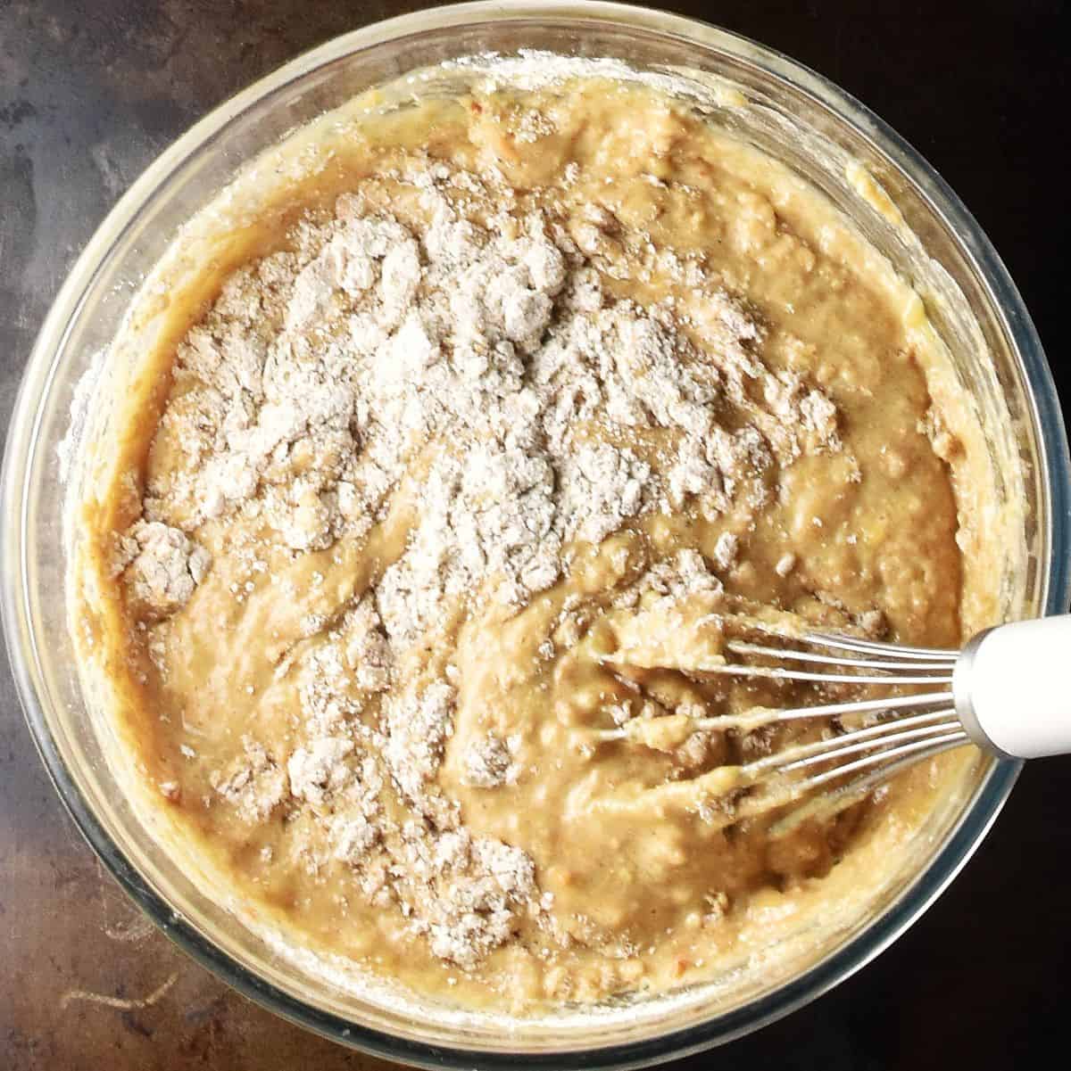 Top down view of creamy peanut butter and banana muffin batter with flour visible in glass bowl with whisk.