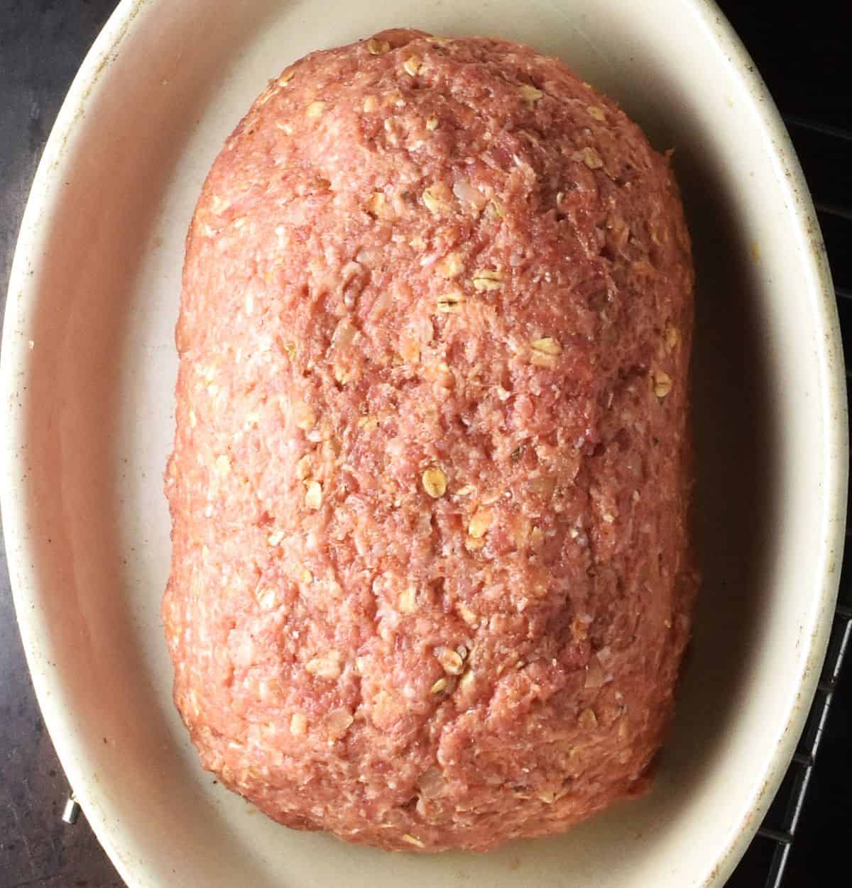 Top down view of unbaked meatloaf with oats in white oval dish.