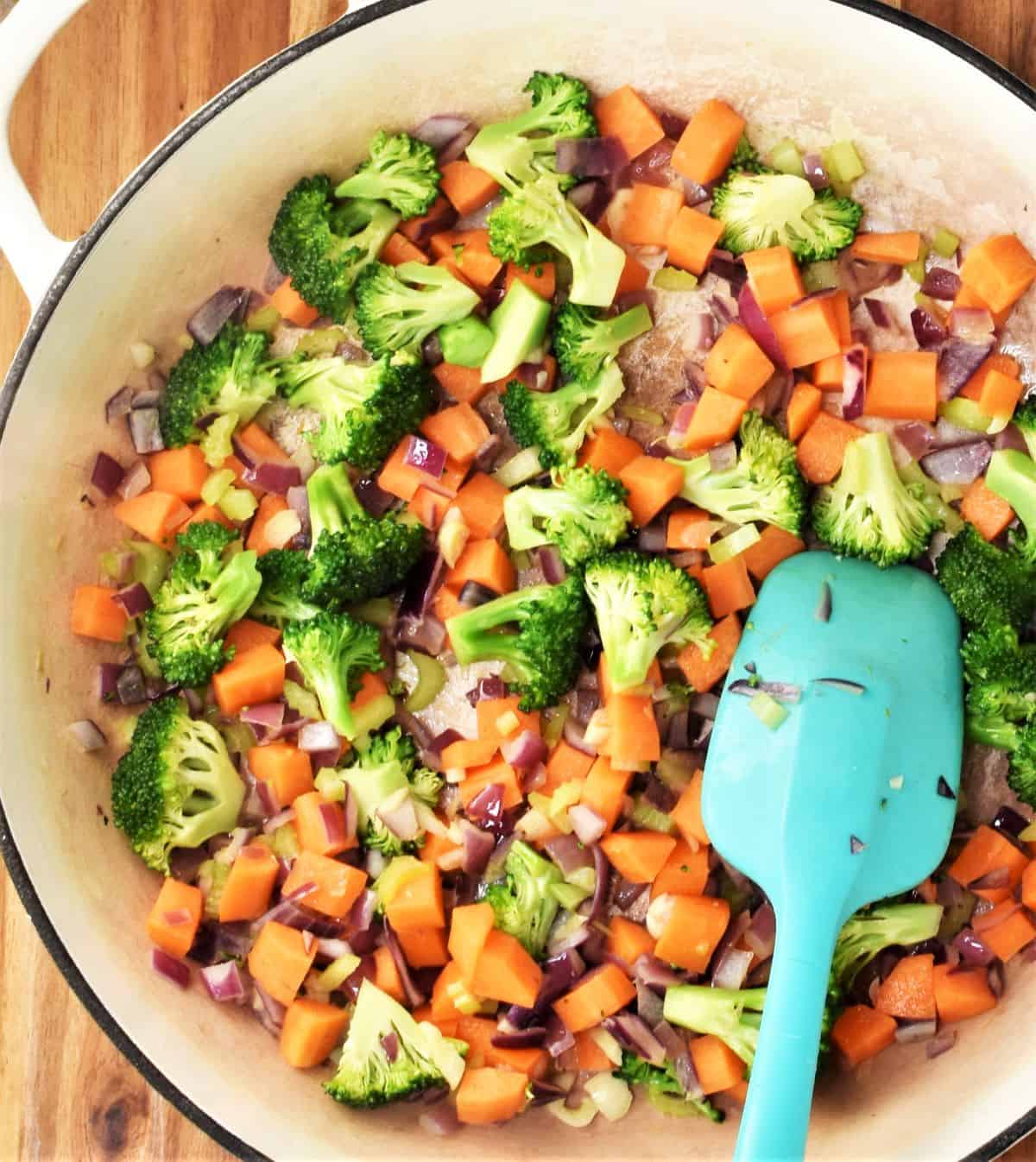 Cooking chopped vegetables in large pan with blue spatula.