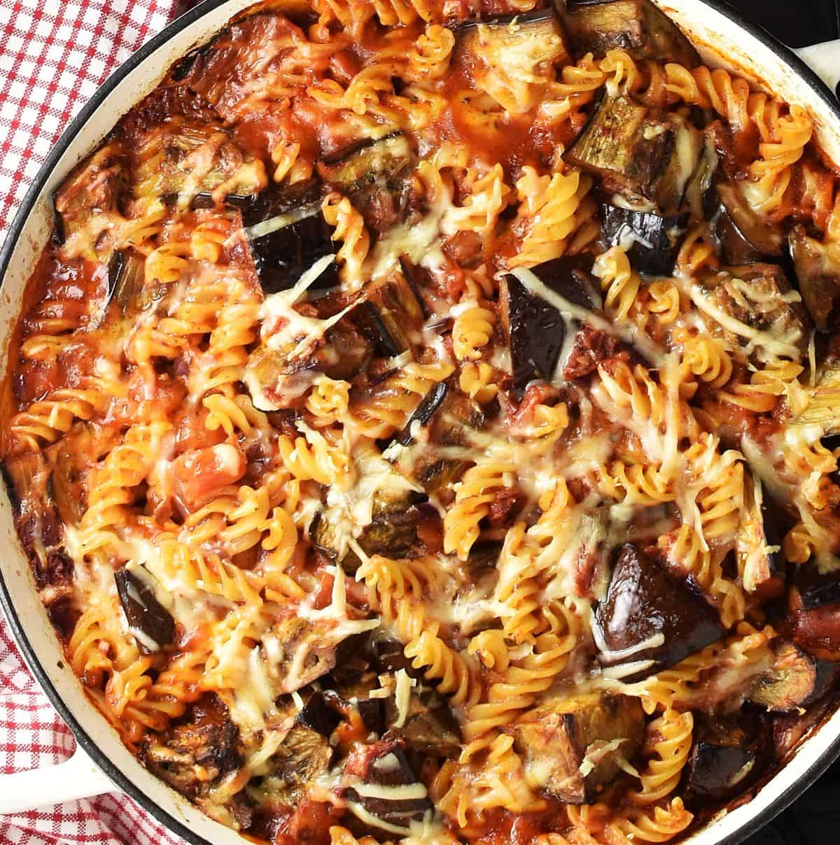 Top down view of baked eggplant pasta in white pan and chequered red cloth in background.