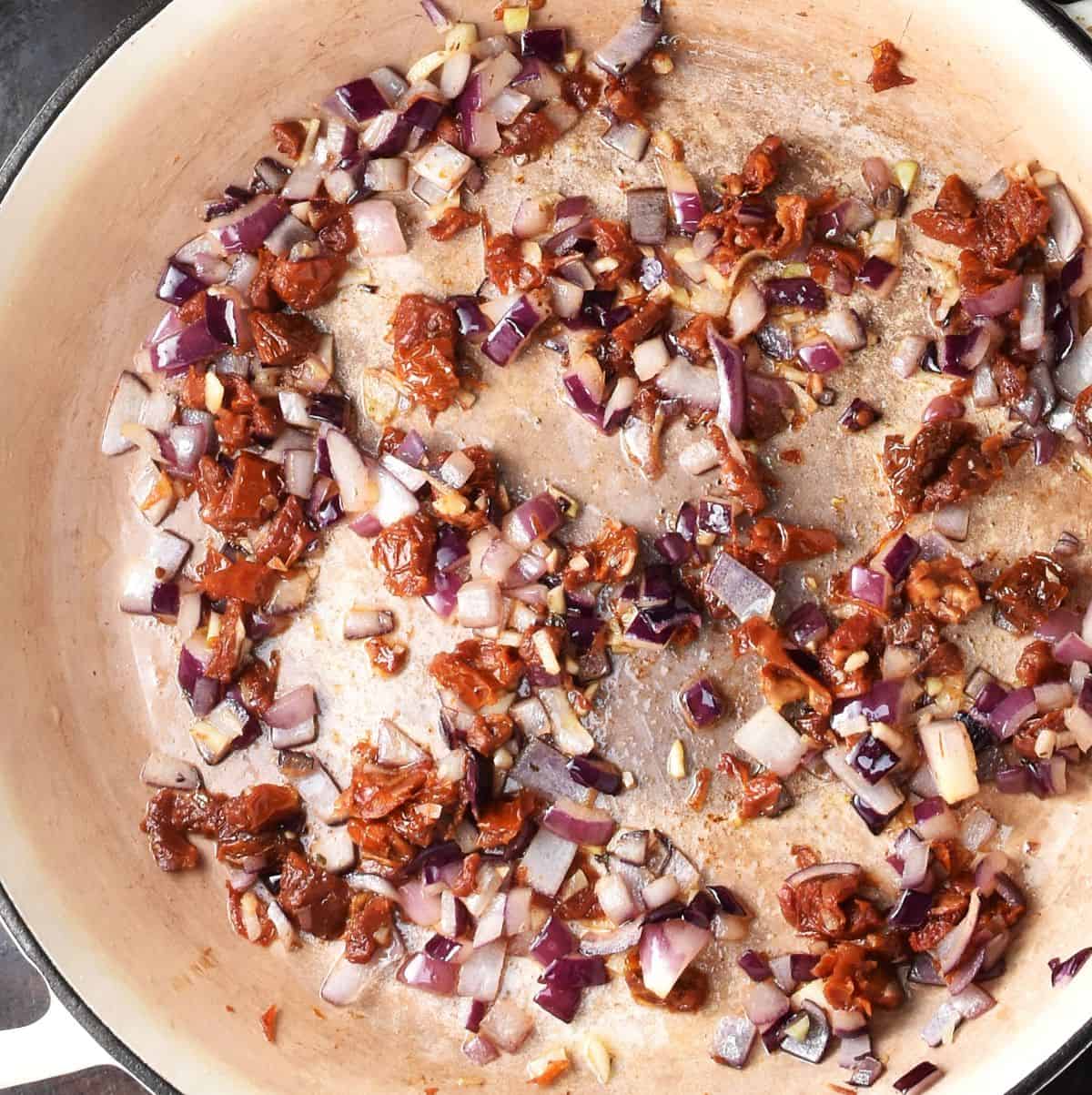 Top down view of chopped red onion and sundried tomatoes in large shallow pan.