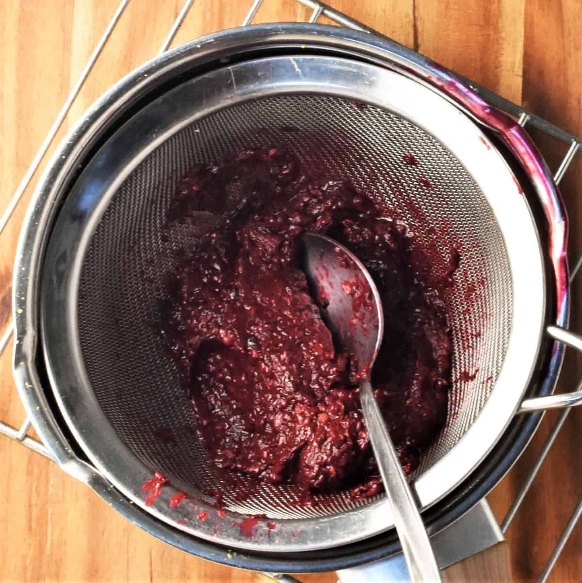 Mashed blackberry mixture in sieve with spoon.