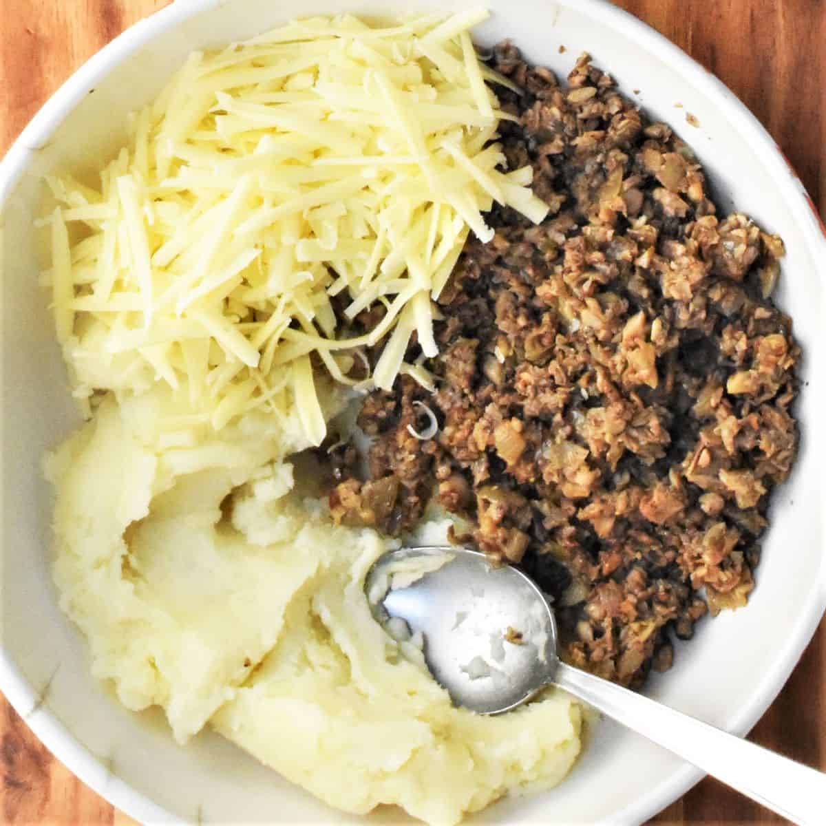 Mashed potato, fried mushroom mixture and grated cheese in white bowl.