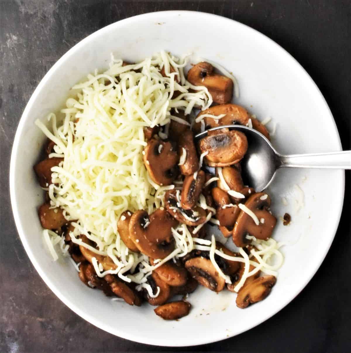 Browned mushroom slices and shredded cheese in white bowl with spoon.