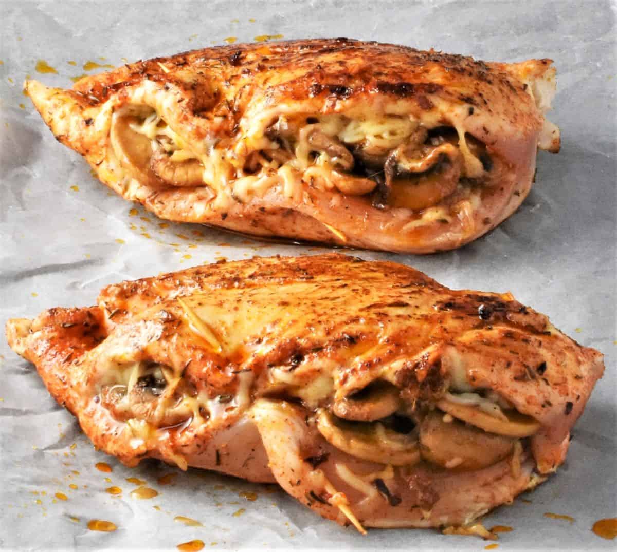 Side view of 2 pre-cooked chicken breasts stuffed with mushrooms and cheese on parchment.