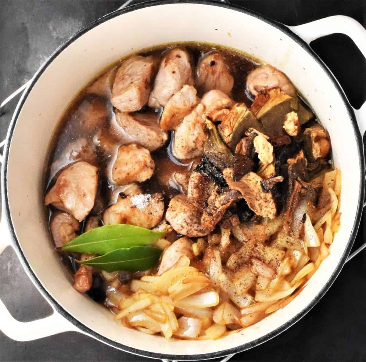 Top down view of browned pork, onion strips and bay leaf with stock in white pot.