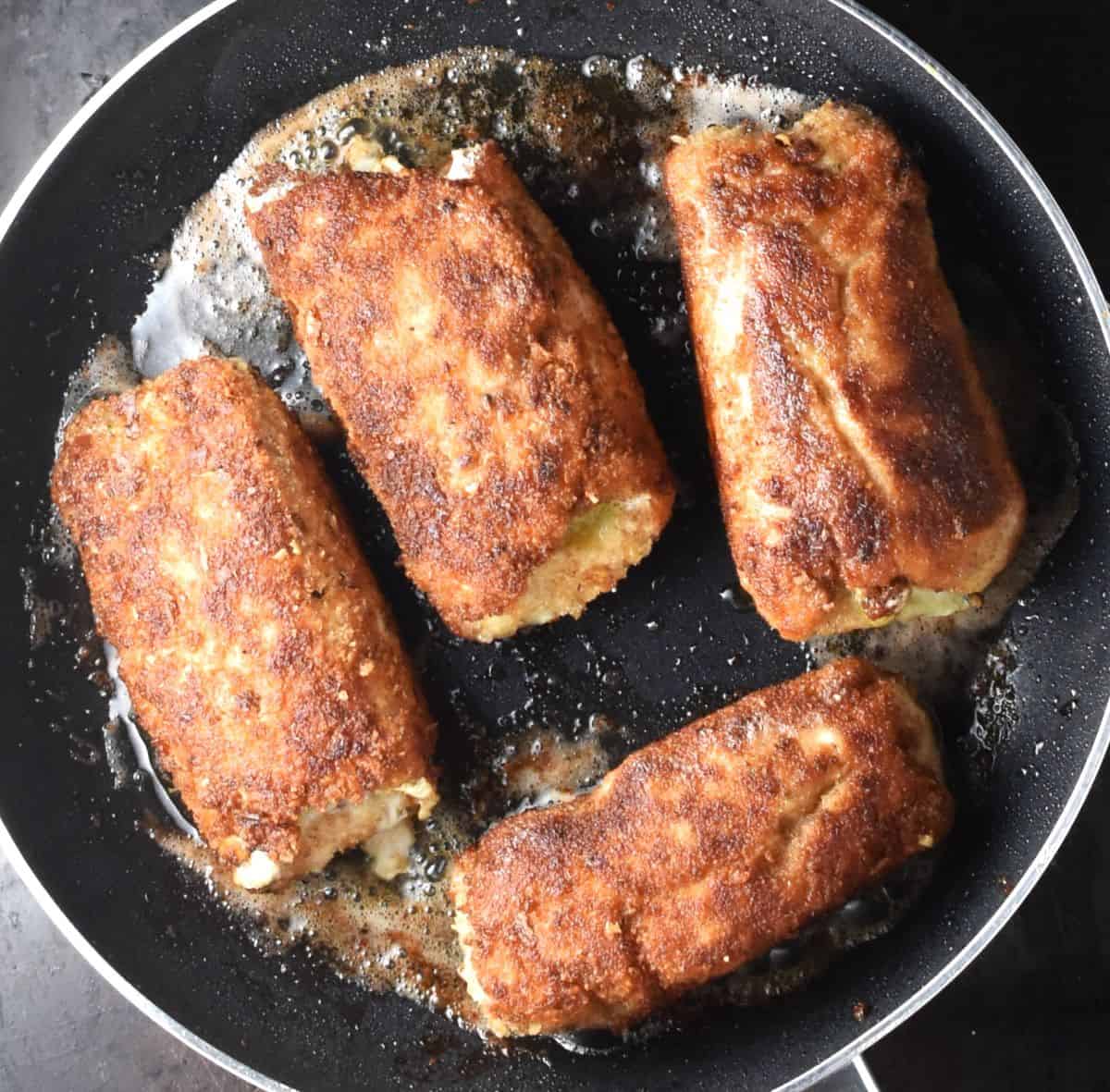 4 breaded rolled up chicken breasts cooking in pan.