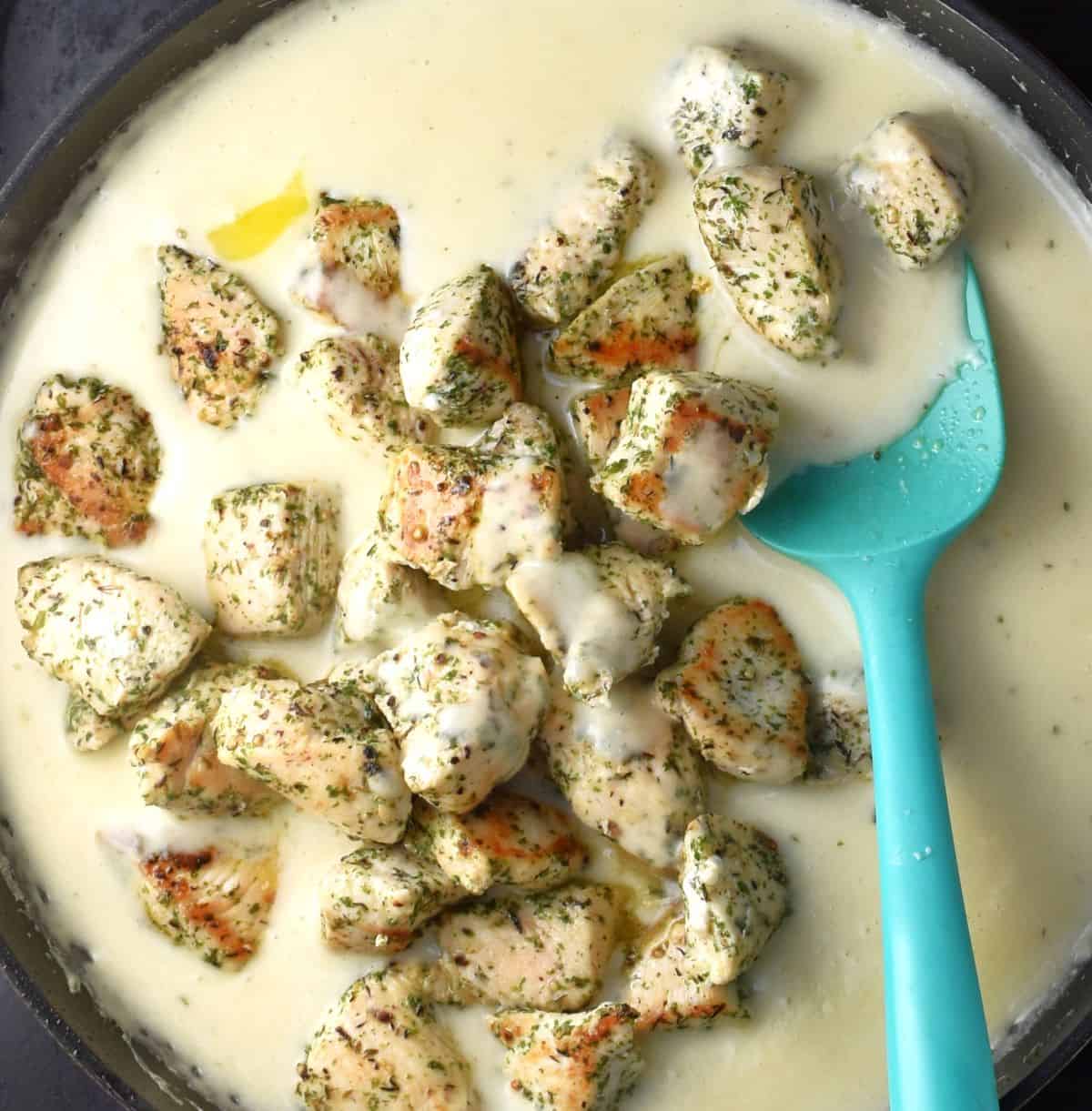 Cooked chicken pieces and creamy sauce in large pan with blue spoon.