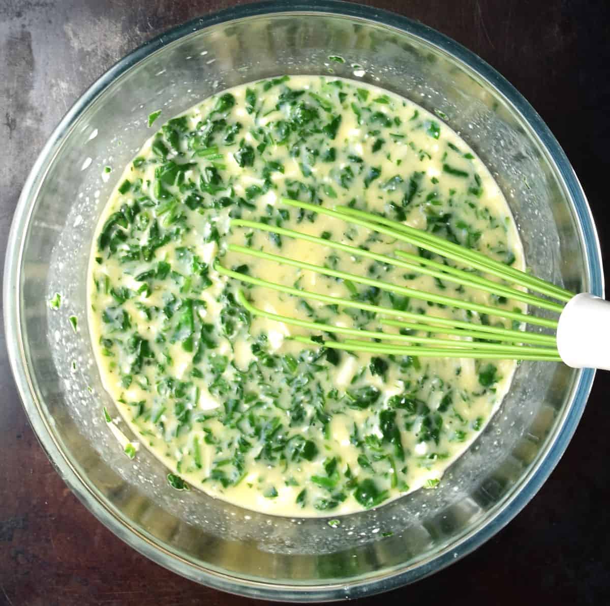 Egg mixture with spinach and whisk in glass bowl.