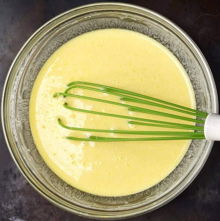 Yellow wet pancake mixture in glass bowl with green whisk.