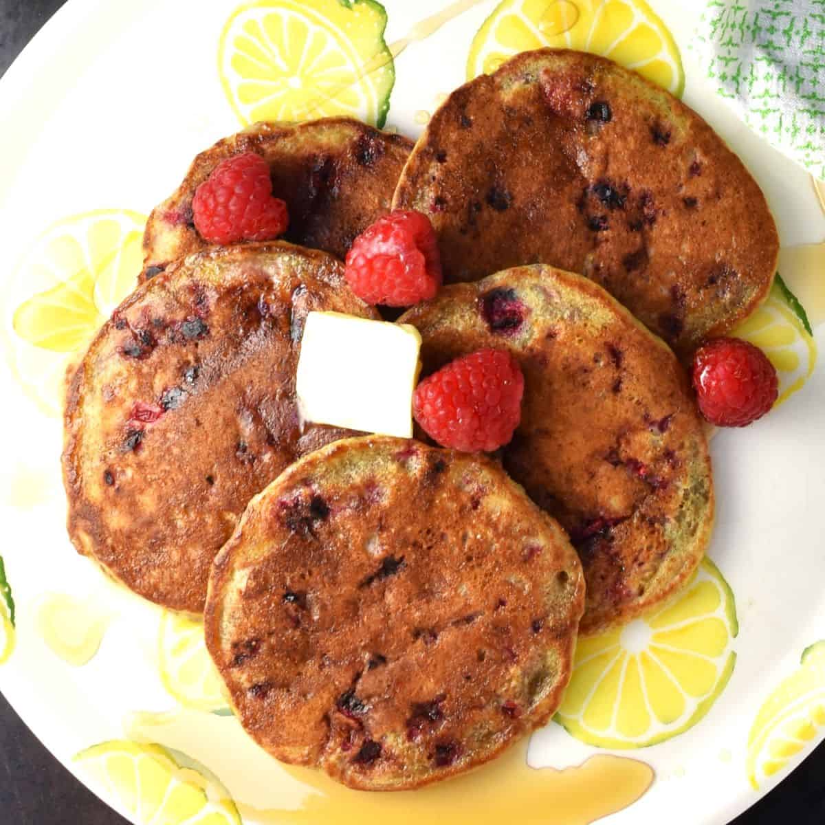 Top down view of 5 raspberry pancakes with fresh raspberries on top of yellow plate.
