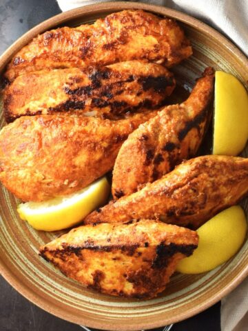 Top down view of browned pieces of yogurt marinated chicken breast with lemon wedges in brown bowl.