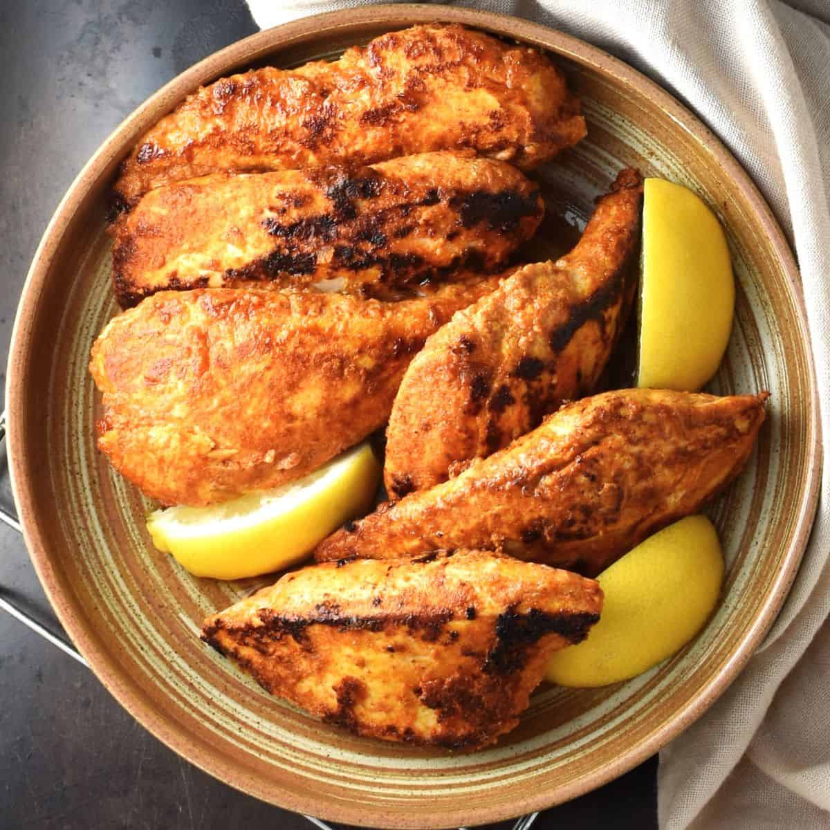 Top down view of browned pieces of yogurt marinated chicken breast with lemon wedges in brown bowl.