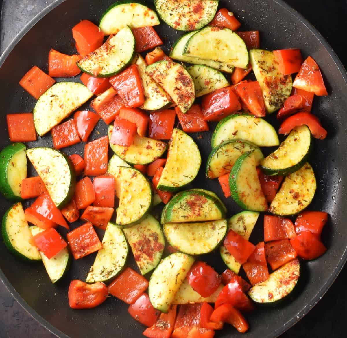 Cooking zucchini and red pepper with herbs in pan.