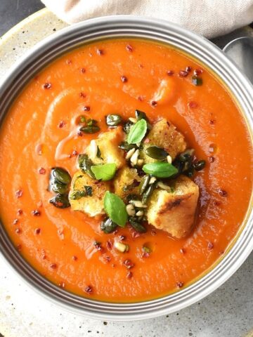 Top down view of creamy butternut squash tomato soup with croutons in grey bowl with spoon on right.