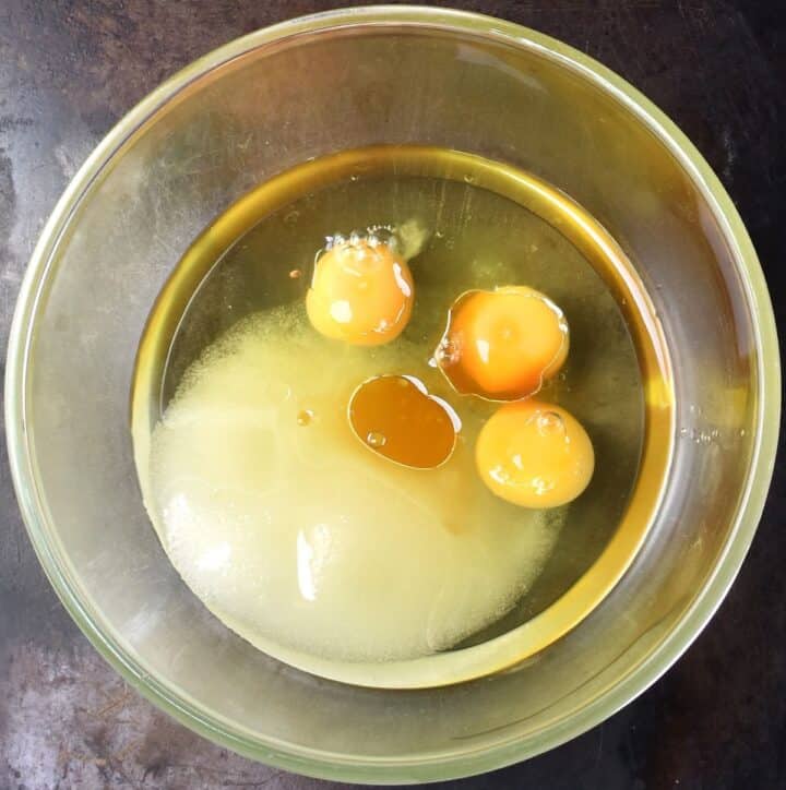 Top down view of 3 eggs with sugar and oil in glass bowl.