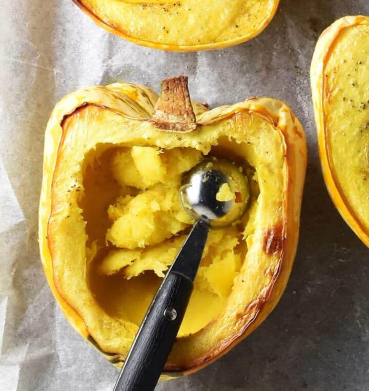 Roasted halved acorn squash hollowed out with melon baller on parchment.