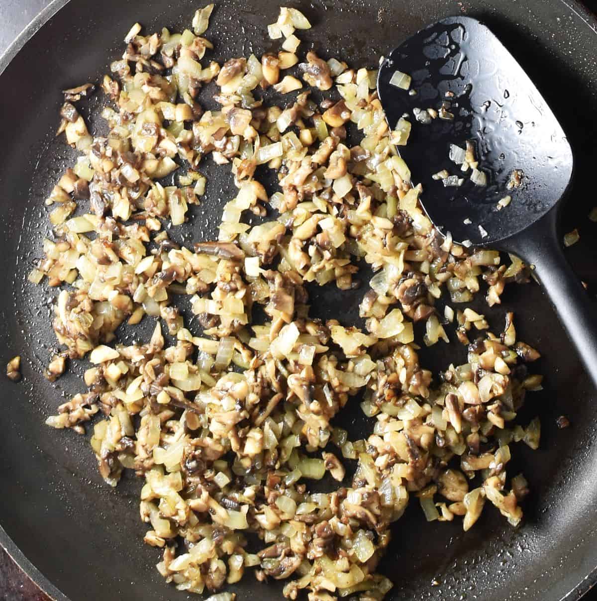 Chopped mushrooms and onion frying in pan with black spoon.