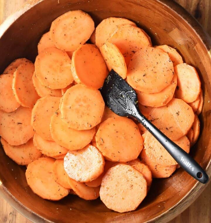 Top down view of thinly sliced sweet potatoes coated in oil and herbs in large wooden bowl with black spatula.