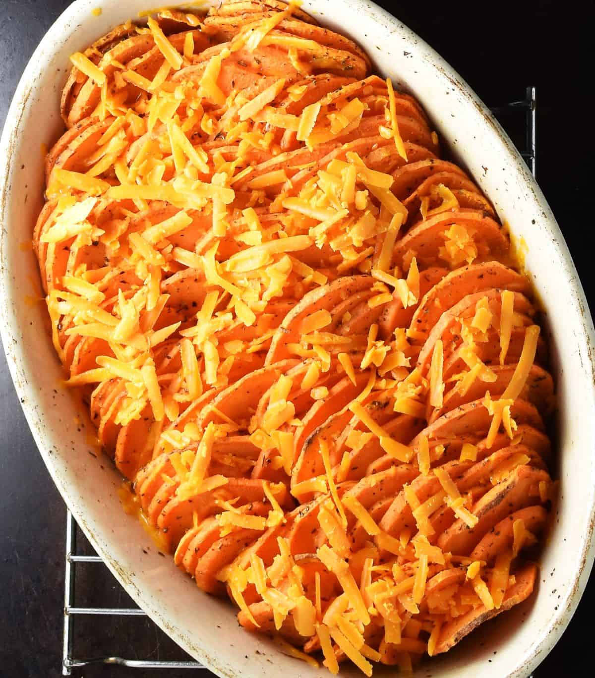 Partly roasted scalloped sweet potatoes with shredded cheese on top in white dish.