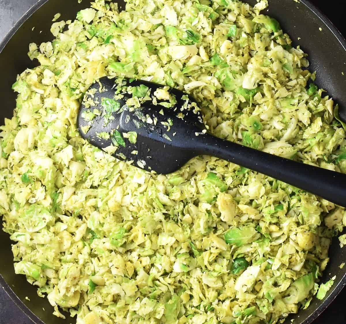 Top down view of shredded sprouts cooking in pan with black spatula.