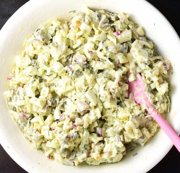 Creamy egg salad wit dill pickles in large white bowl with pink spatula.