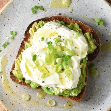 Top down view of avocado feta toast with scallions and honey on speckled plate.