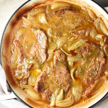 Top down view of braised pork steaks with onion in large shallow pan.