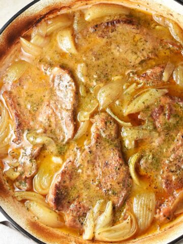 Top down view of braised pork steaks with onion in large shallow pan.
