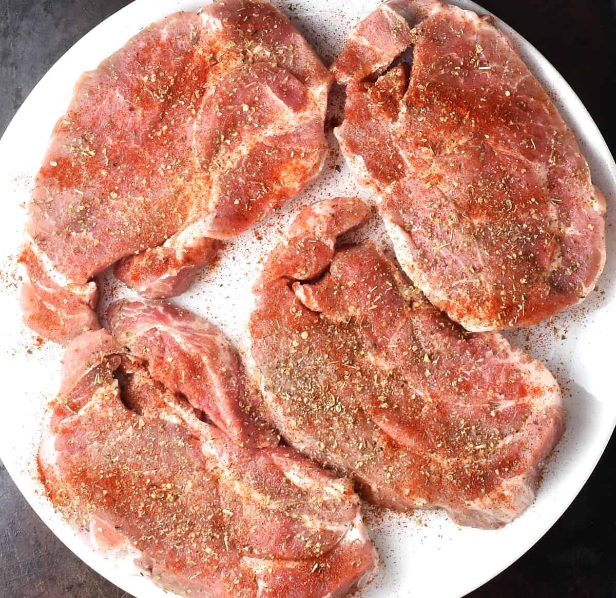 Seasoned raw pork steaks on white plate.