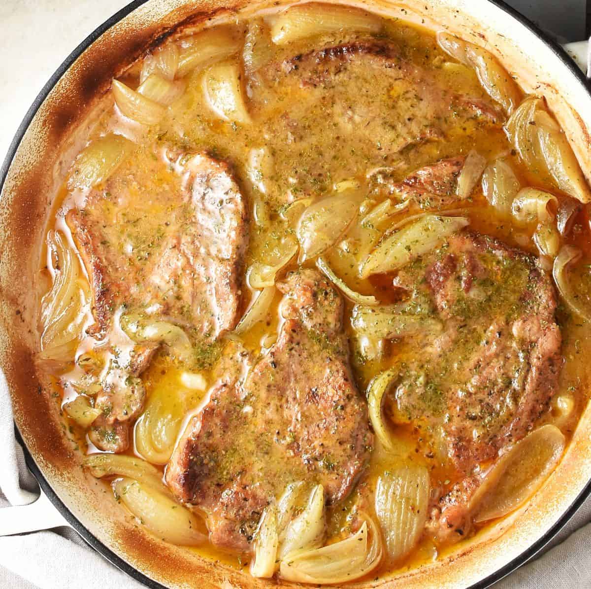 Top down view of 4 pork steaks with onion in sauce in white shallow pan.