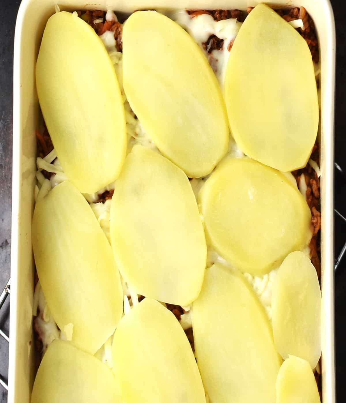 Sliced of potato arranged on top of meat layer in rectangular lasagna dish.