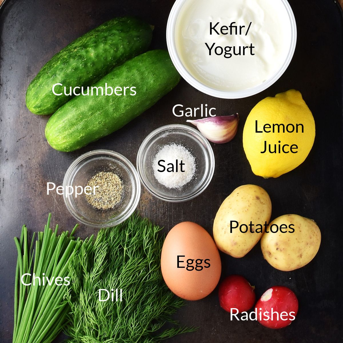 Top down view of ingredients for making traditional Polish cold cucumber soup on top of dark surface.