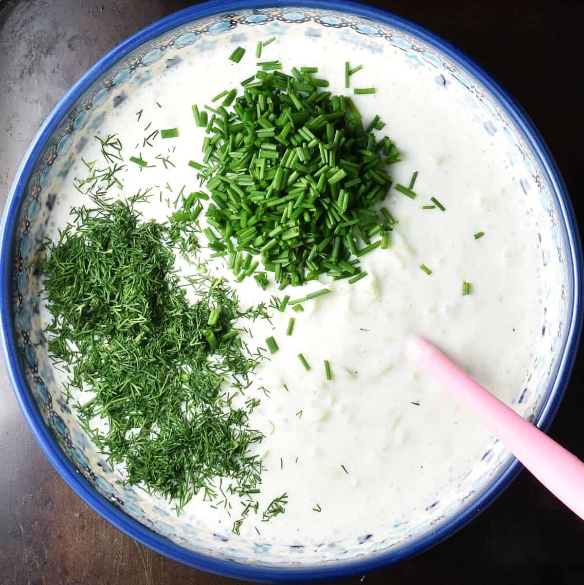 Top down view of creamy cold cucumber soup with dill and chives in blue bowl with pink spoon.