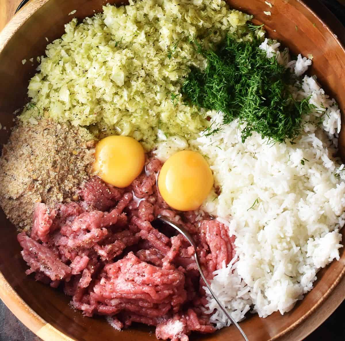 Top down view of ground meat, shredded cabbage, rice, 2 raw eggs, breadcrumbs and herbs in brown bowl.