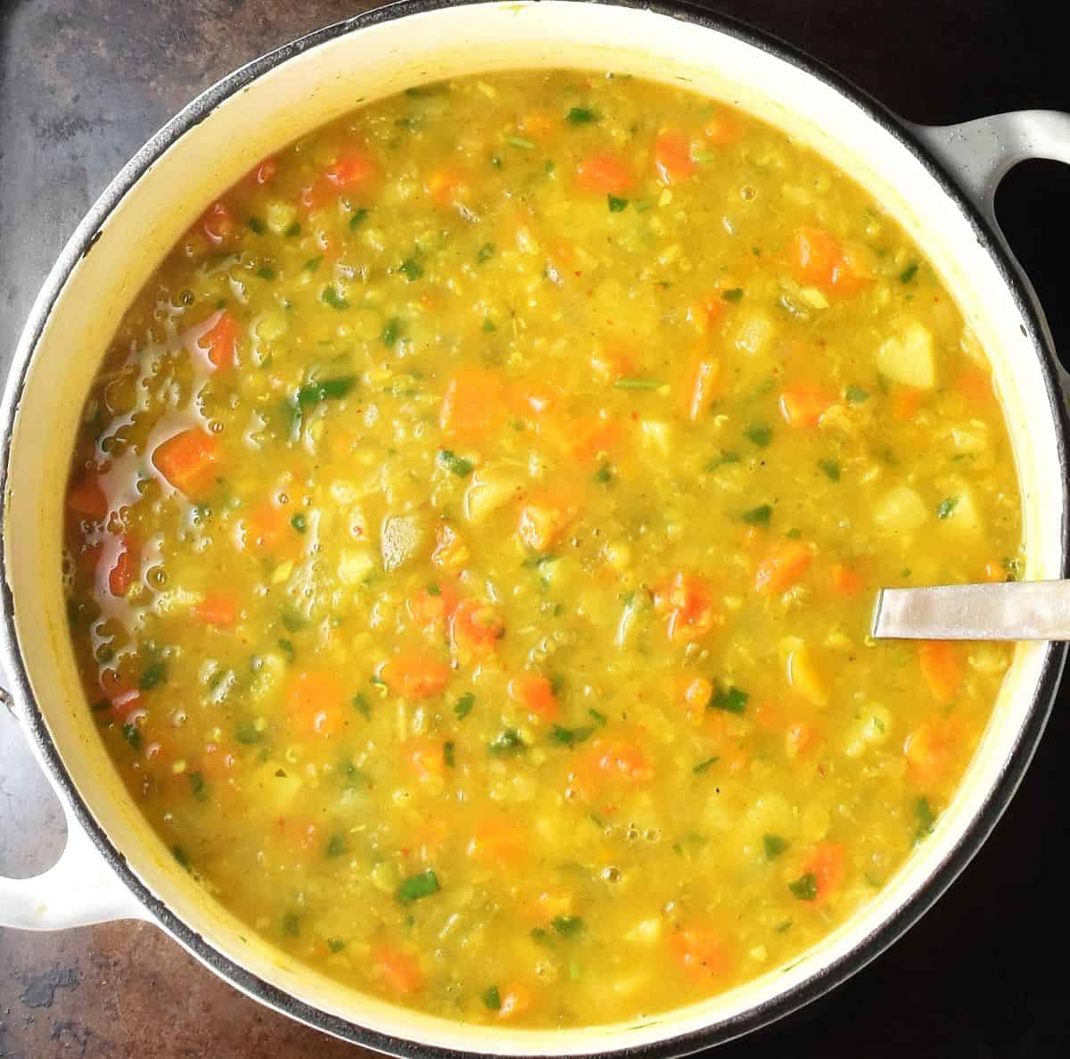 Top down view of chunky mulligatawny soup with vegetables and lentils in white pot with ladle.