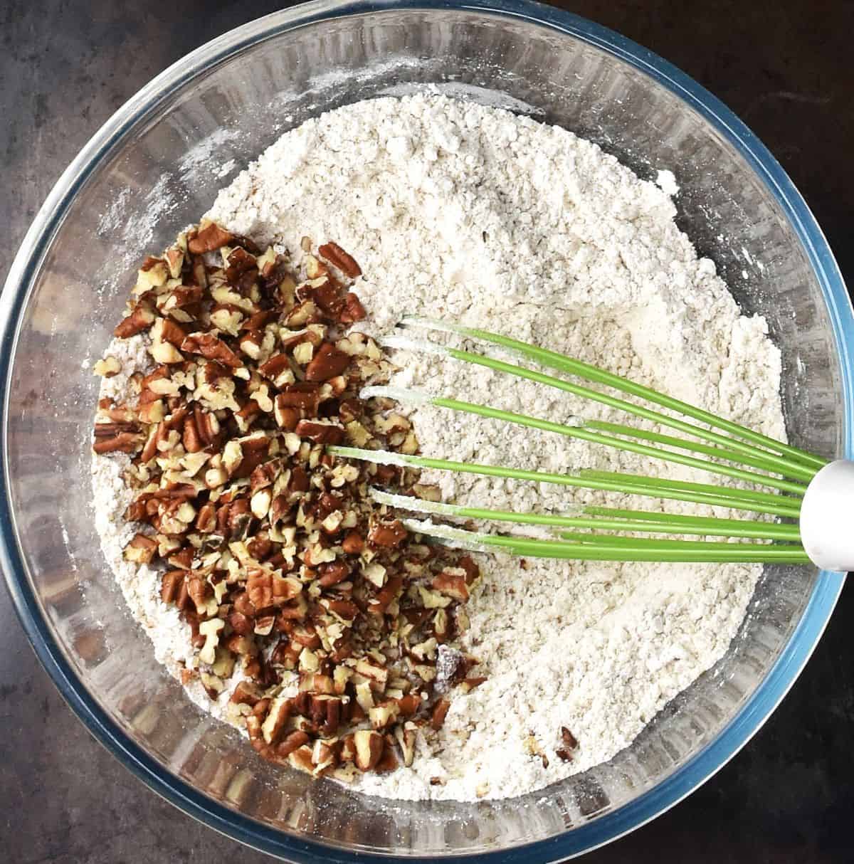 Top down view of dry muffin mixture with chopped pecans and green whisk.