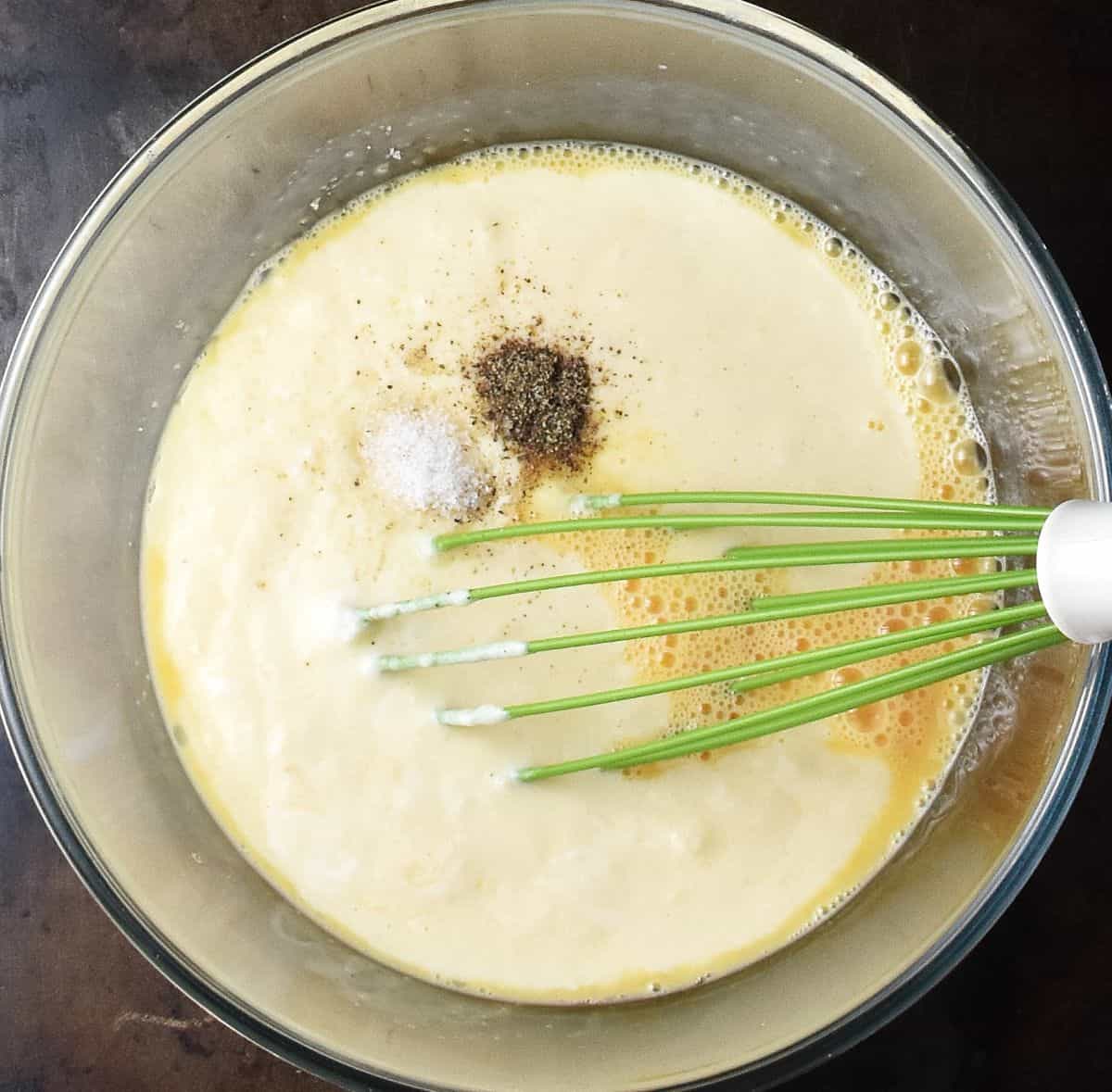 Creamy egg potato mixture with salt and pepper in glass bowl with whisk.