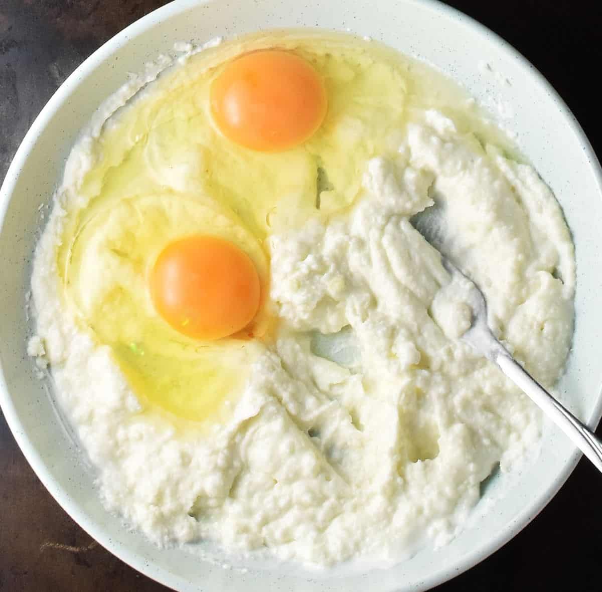 Creamy potato mixture with 2 raw eggs in shallow bowl with fork.