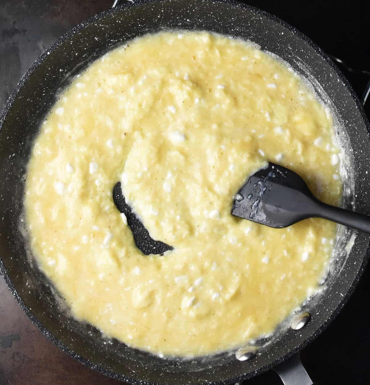 Cooking scrambled eggs with cottage cheese in black pan with black spatula.