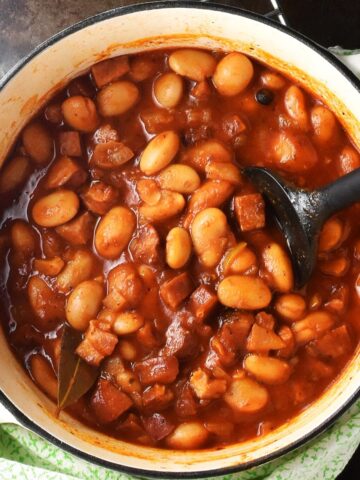 Top down view of Polish beans in tomato sauce in white pot with black spoon.