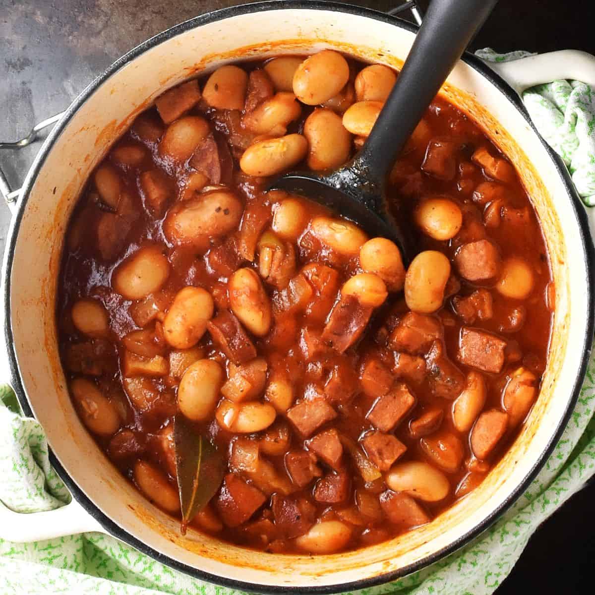 Lima beans in thick tomato sauce with sausage in white pot with black spatula.