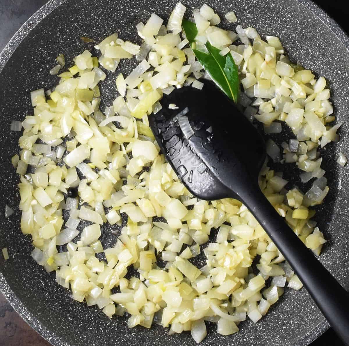 Top down view of chopped sauteed onion with bay leaf in large pan with black spatula.