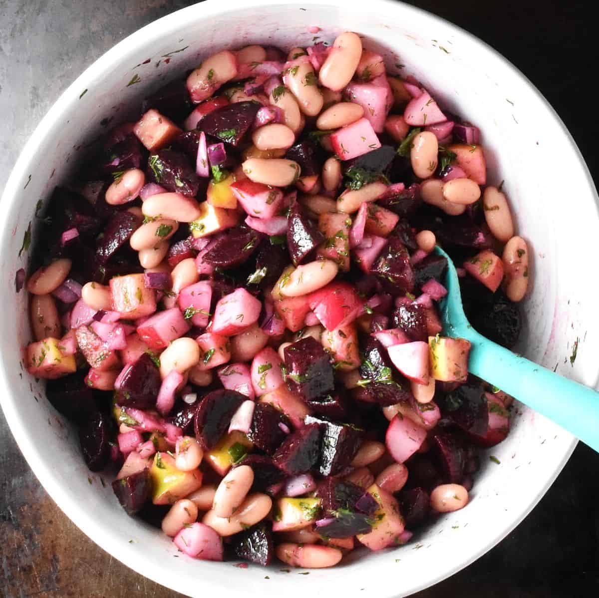 Stirred pickled beet salad with beans in white bowl with blue spoon.
